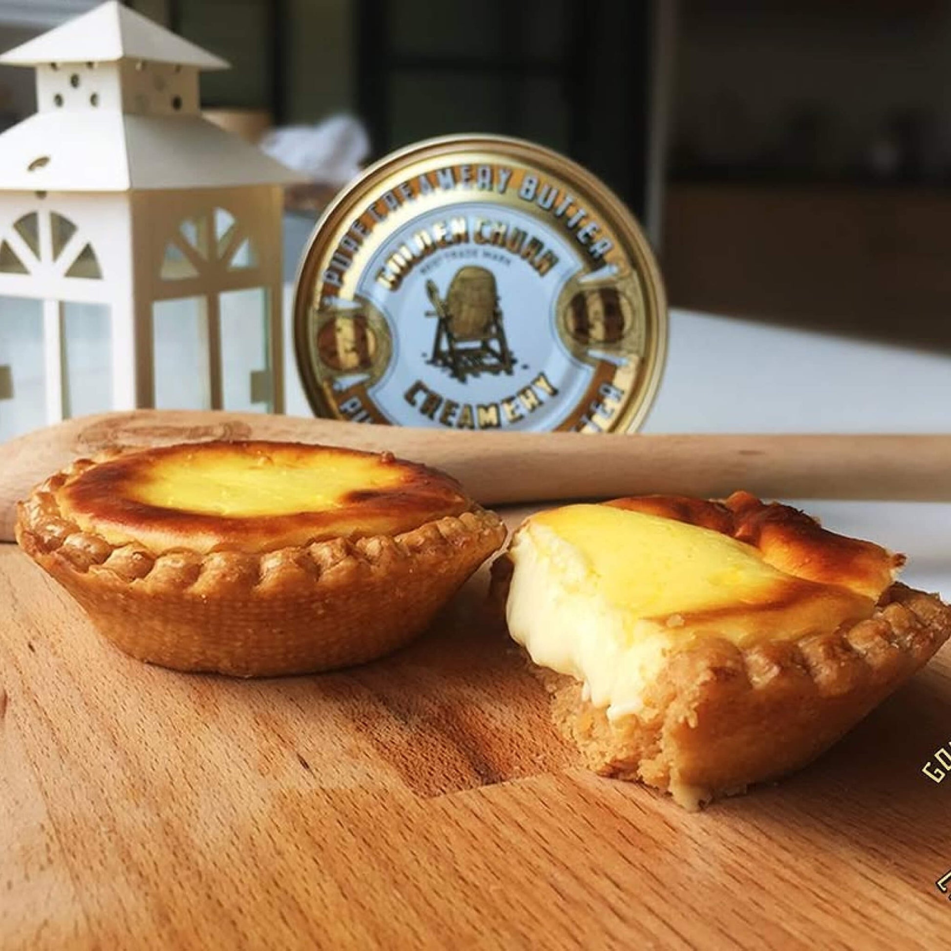Two egg tarts on a wooden board with a tin of golden churn butter in the background.