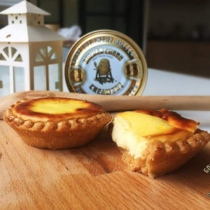 Two egg tarts on a wooden board with a tin of golden churn butter in the background.